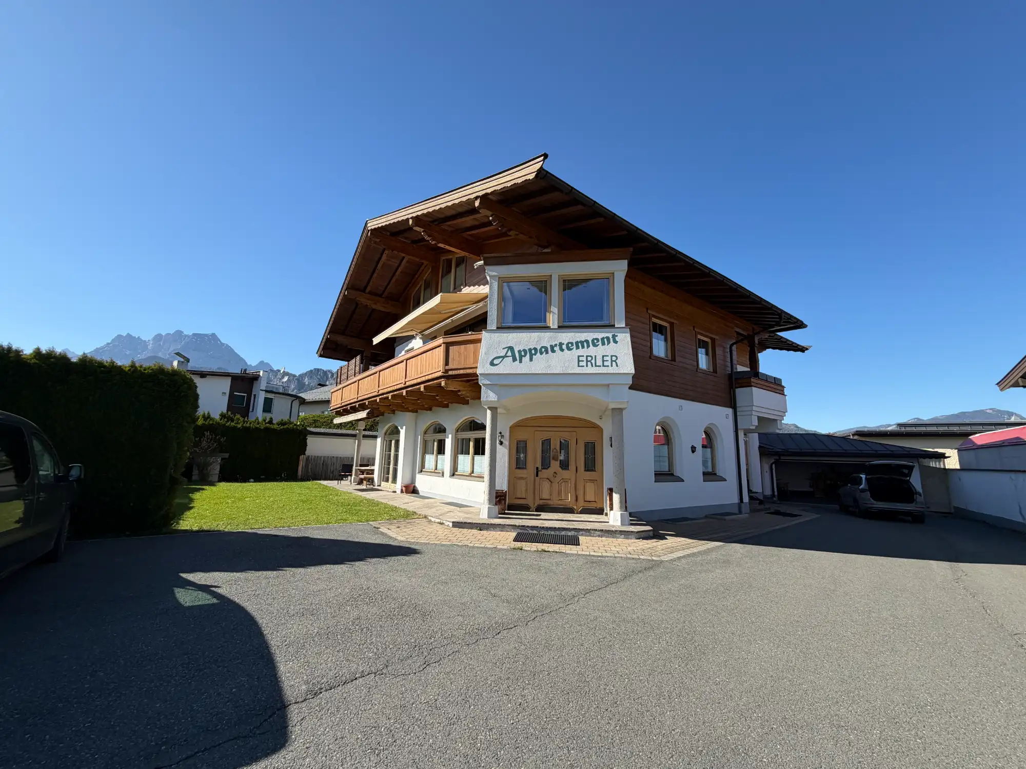 Appartement Erler in St. Johann in Tirol – Außenansicht des Hauses mit Holzbalkonen und Blick auf den Wilden Kaiser bei Sonnenschein.