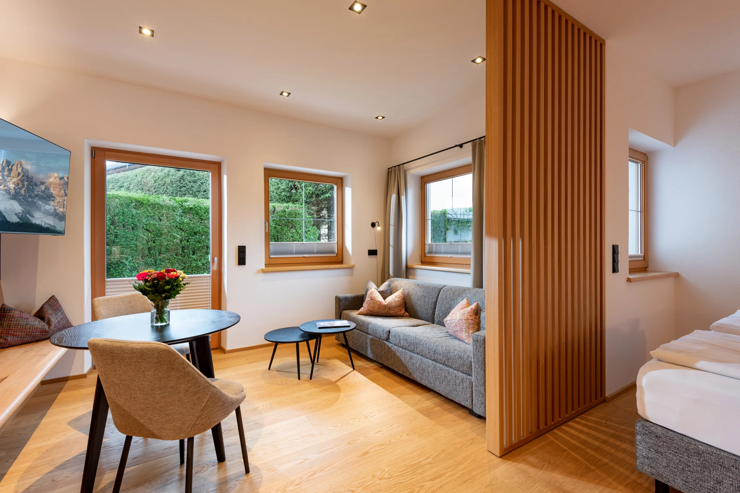 Wohn- und Schlafbereich mit grauer Couch, Holzlamellen-Raumteiler und Gartenblick im Studio mit Garten des Appartement Erler.