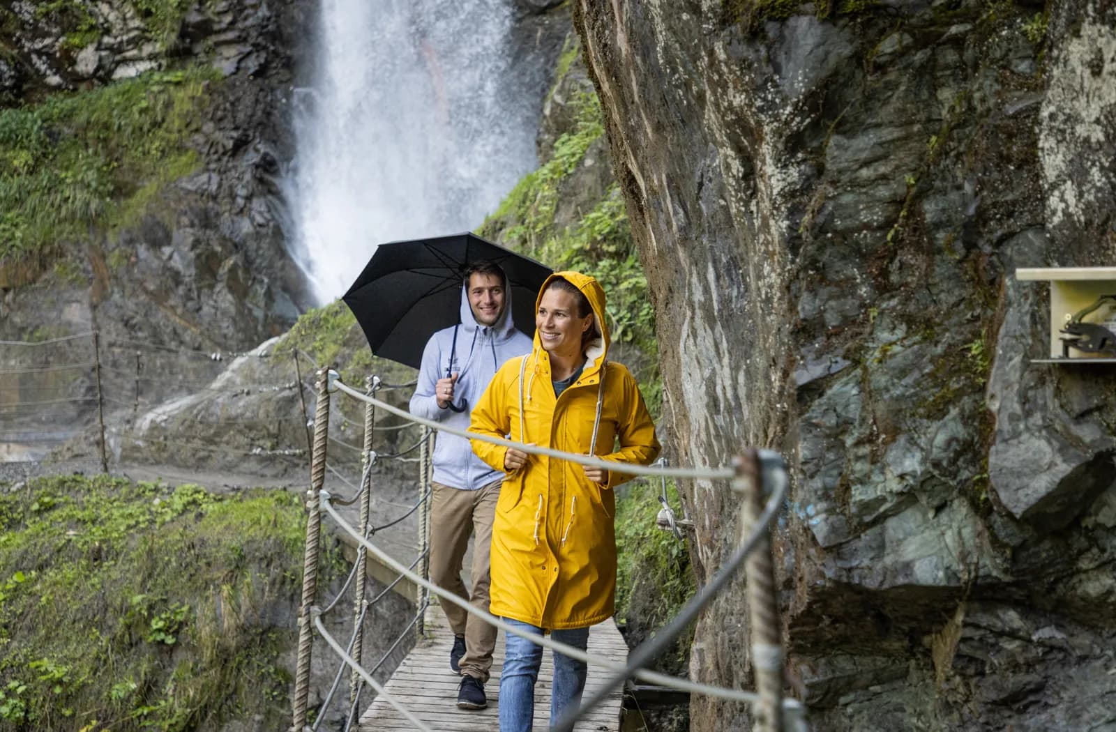 Zwei Wanderer mit Regenschirm und gelber Regenjacke auf dem Steg am Eifersbacher Wasserfall bei St. Johann in Tirol.