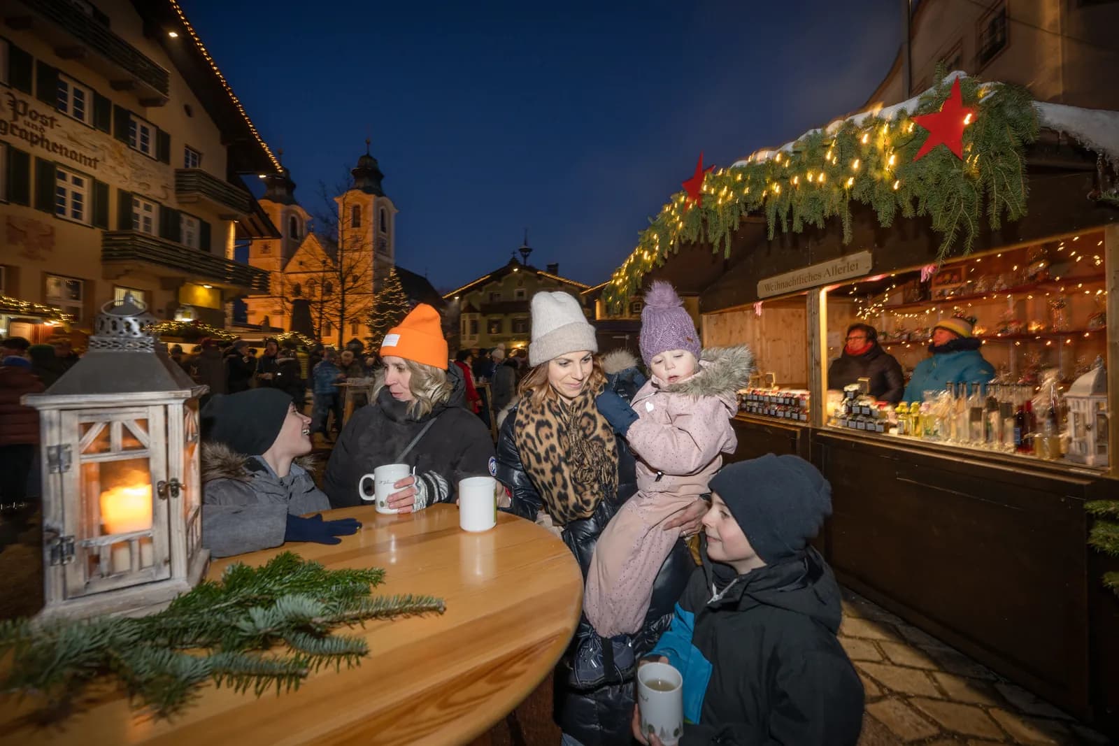 Familie mit dampfenden Tassen an einem Stehtisch vor einem beleuchteten Stand des St. Johanner Weihnachtsmarkts; im Hintergrund die Dekanatspfarrkirche und ein Christbaum am Hauptplatz.
