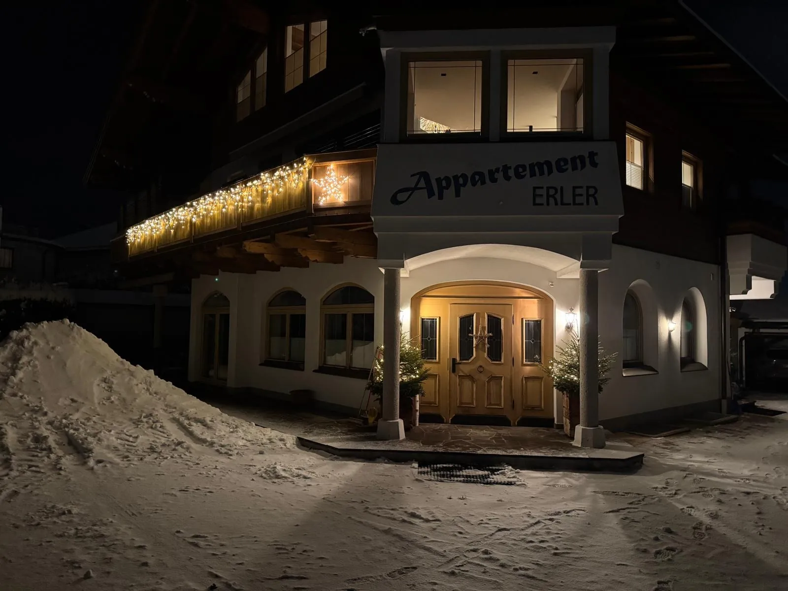Das Appartement Erler am Abend: beleuchteter Eingang und Lichterkette am Balkon über dem verschneiten Vorplatz.