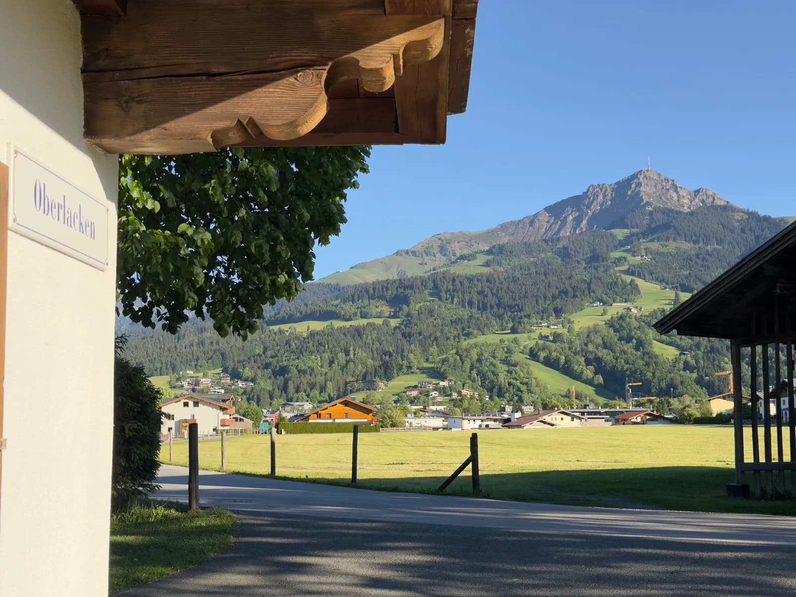 Das Hausschild „Oberlacken“ unter dem geschnitzten Dachbalken, dahinter Wiesen und das Kitzbüheler Horn im Sommer.