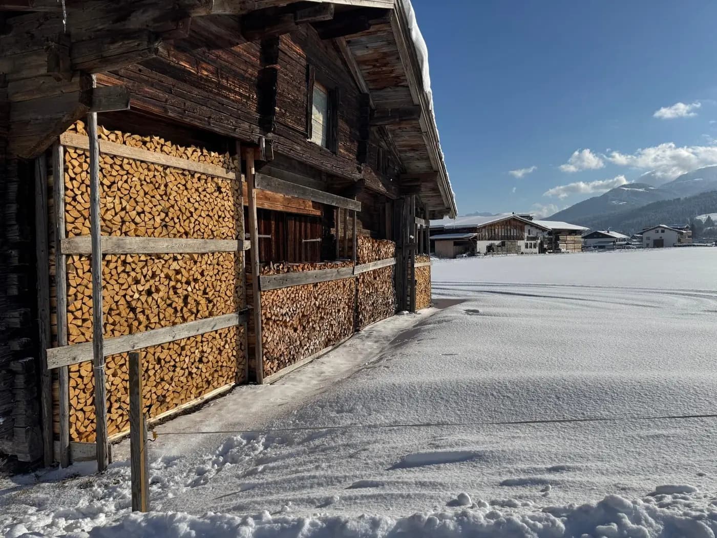 Aufgeschichtetes Brennholz an der Holzwand des Stadels am Bauernhof Oberlacken, davor frischer Schnee.